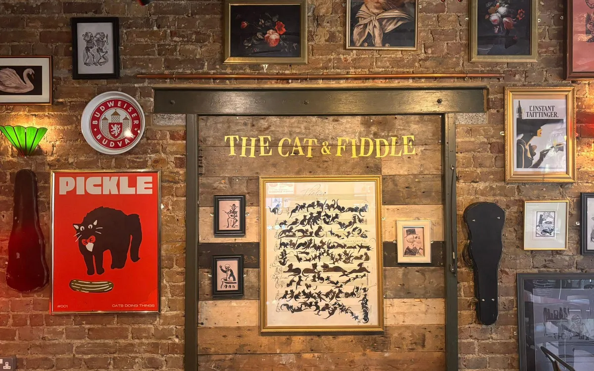 Cat & Fiddle’s brick back wall, covered in framed prints and gig posters, with its name painted in gold